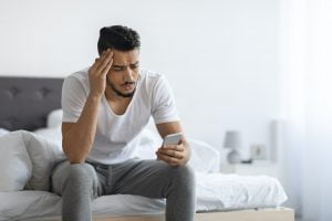 Bad News. Upset Arab Man Sitting On Bed And Looking At Smartphone